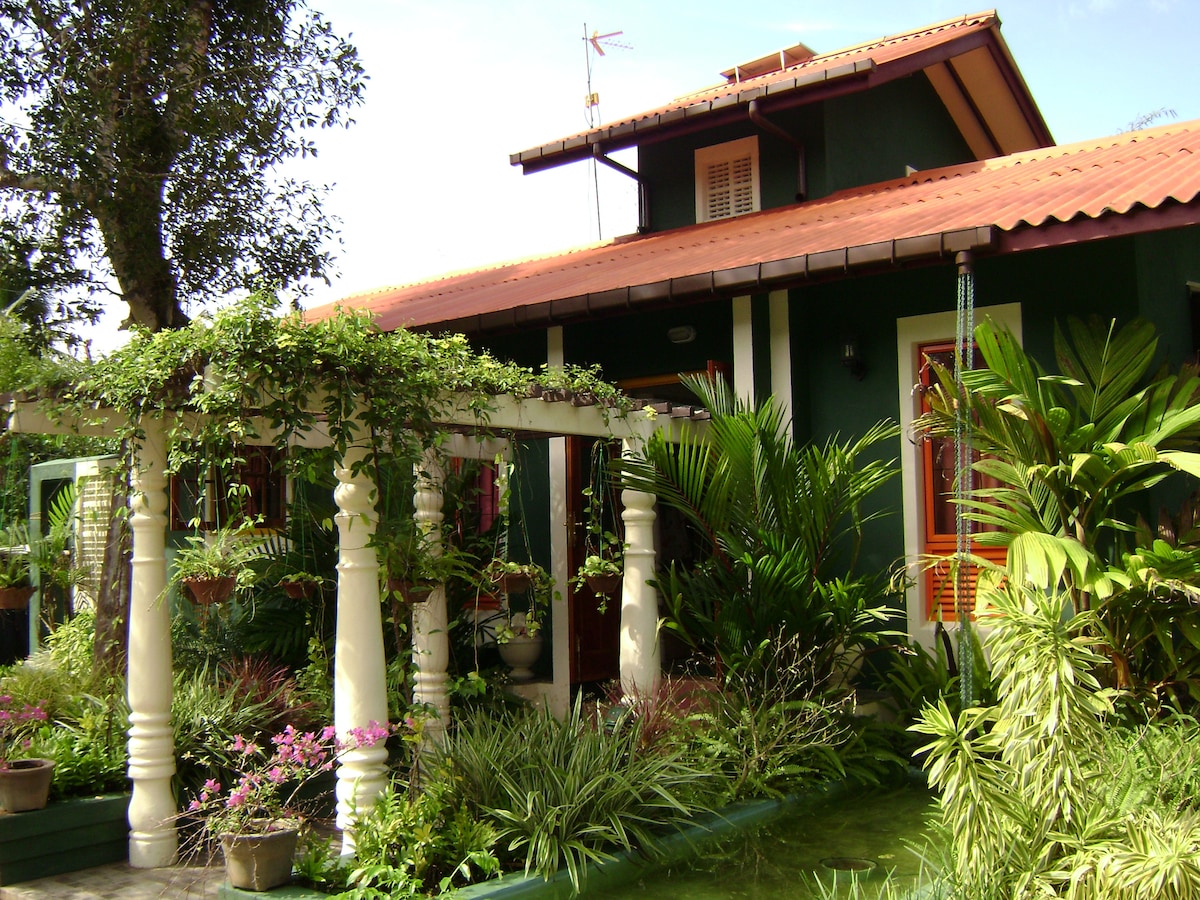 The bungalow is surrounded by lush greenery, featuring a covered porch with intricately carved columns. Tropical plants and flowers are positioned throughout, creating a vibrant entrance. A shallow pond runs alongside, enhancing the natural aesthetic of the outdoor space.