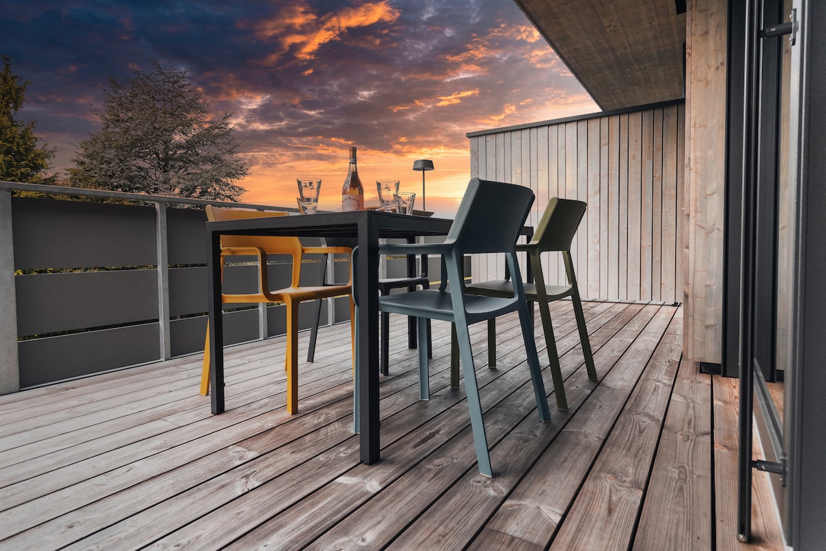 A spacious balcony features a wooden deck with a table and four chairs in varied colors. The setting is illuminated by the warm colors of a sunset, providing a serene backdrop for relaxation or dining outdoors.