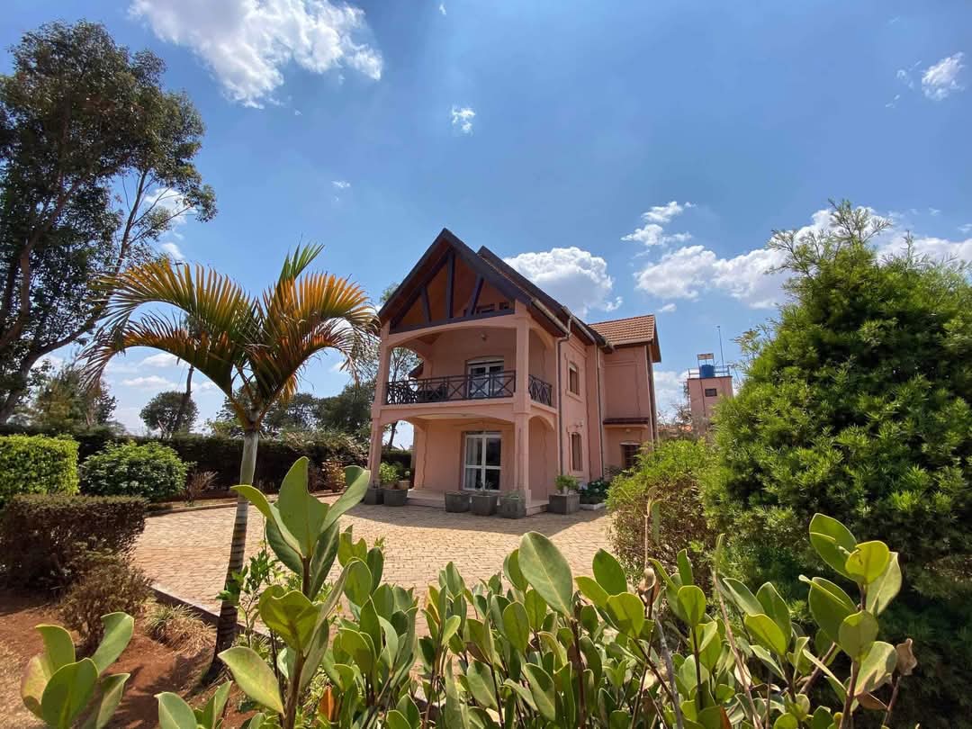 A two-story pink house is shown, featuring a distinct roofline and a spacious balcony. Surrounding greenery includes well-maintained shrubs and palm plants. A clear blue sky with scattered clouds creates an open and airy ambiance.