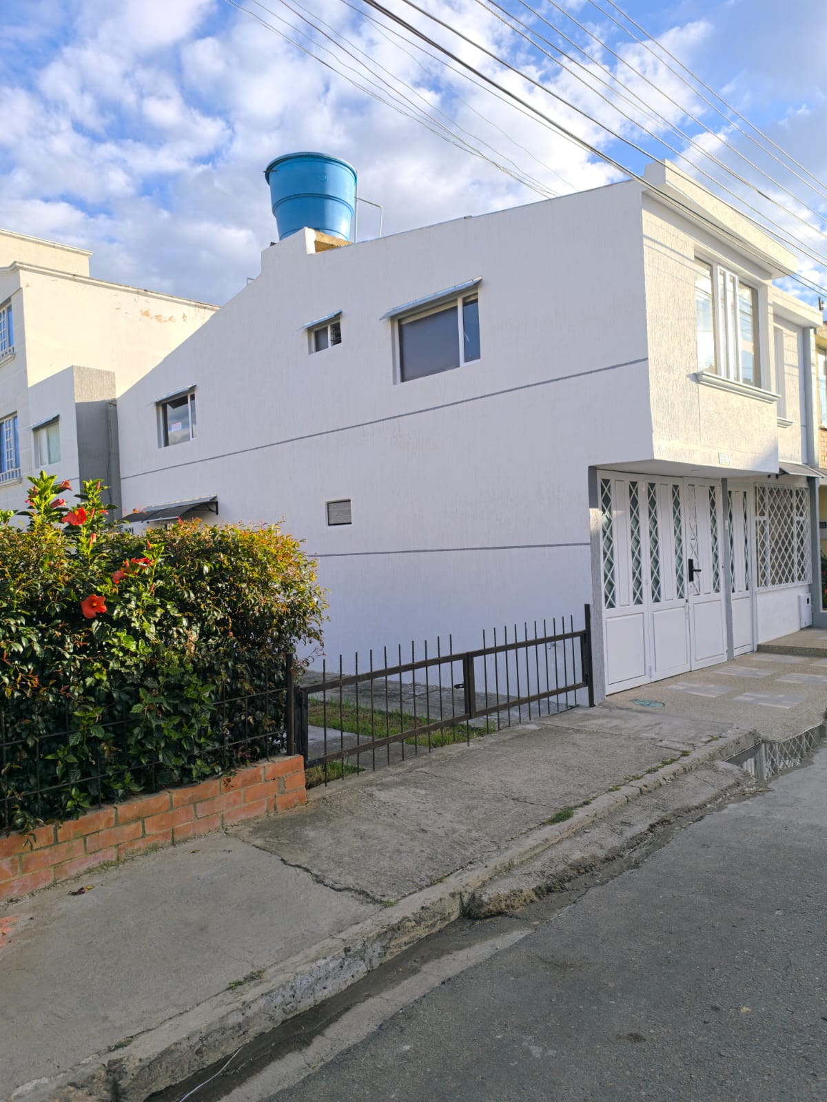 The exterior of a white building is featured, showcasing a well-maintained facade with large windows. A metal gate in front adds security. A small garden with colorful flowers is visible alongside the pathway, creating a welcoming entrance to the property.