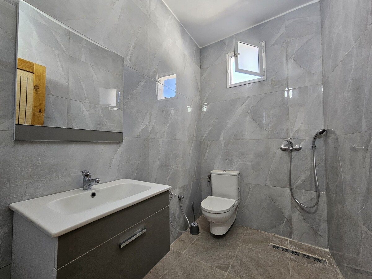 The bathroom features contemporary gray tiles that cover the walls and floor. A modern vanity with a sink is positioned against one wall, and a large mirror reflects the space. Natural light enters through a small window, while a shower area and toilet complete the layout.