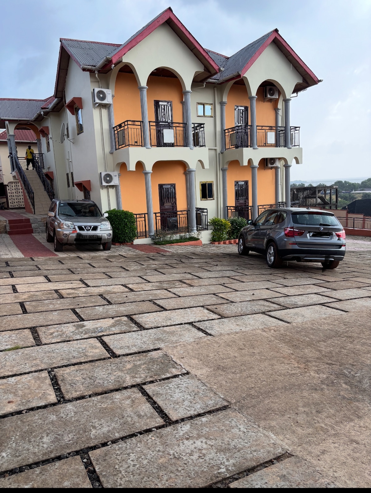 A two-story house is depicted, featuring arched windows and balconies on the upper level. The exterior is painted in warm tones, complemented by neatly arranged plants. A paved driveway accommodates two parked vehicles, while a staircase leads to the entrance.