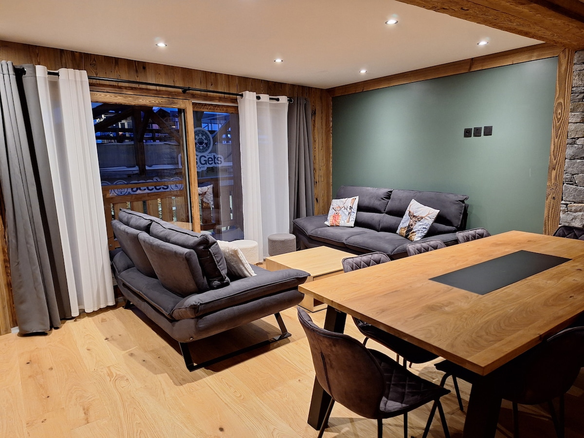 A spacious living area features comfortable seating with dark sofas positioned around a wooden dining table. Large windows allow natural light to fill the space, with soft curtains framing the view. Rustic wood accents contribute to the overall design.