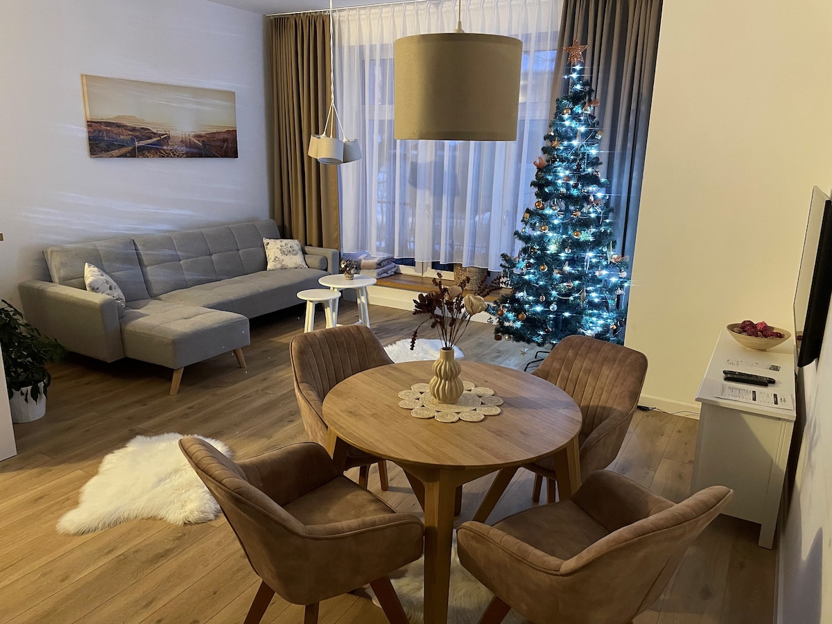 A living area features a cozy grey sofa and a round wooden dining table surrounded by four upholstered chairs. A decorative tree adorned with lights stands nearby, and large windows allow natural light to fill the space, complemented by soft, neutral-toned curtains.