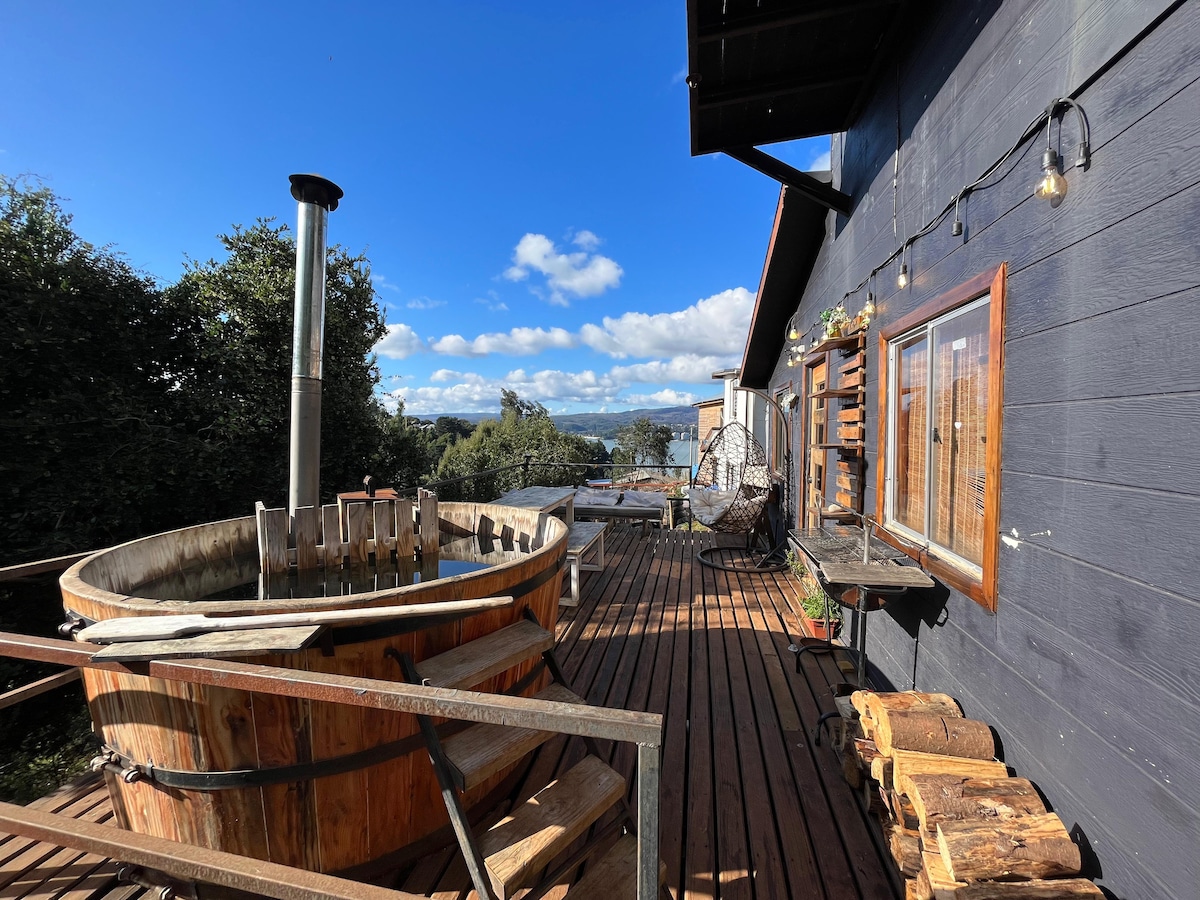 An outdoor terrace is shown featuring a wooden hot tub positioned beside a wooden deck. Views of surrounding nature and hills are visible in the background, complemented by a blue sky with scattered clouds. Warm lighting from overhead fixtures enhances the inviting space.