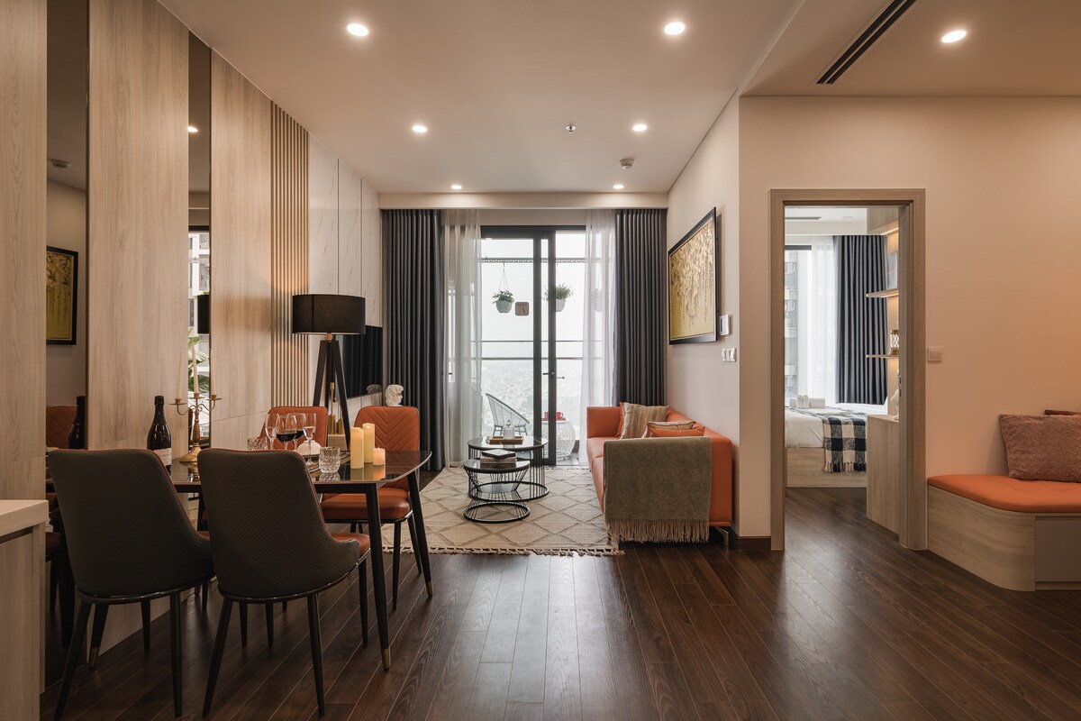 The living area features a dining table set for four, complemented by stylish chairs. A cozy sofa invites relaxation, while large glass doors lead to a balcony. Natural light fills the space, enhancing the warm wooden floors and contemporary decor.