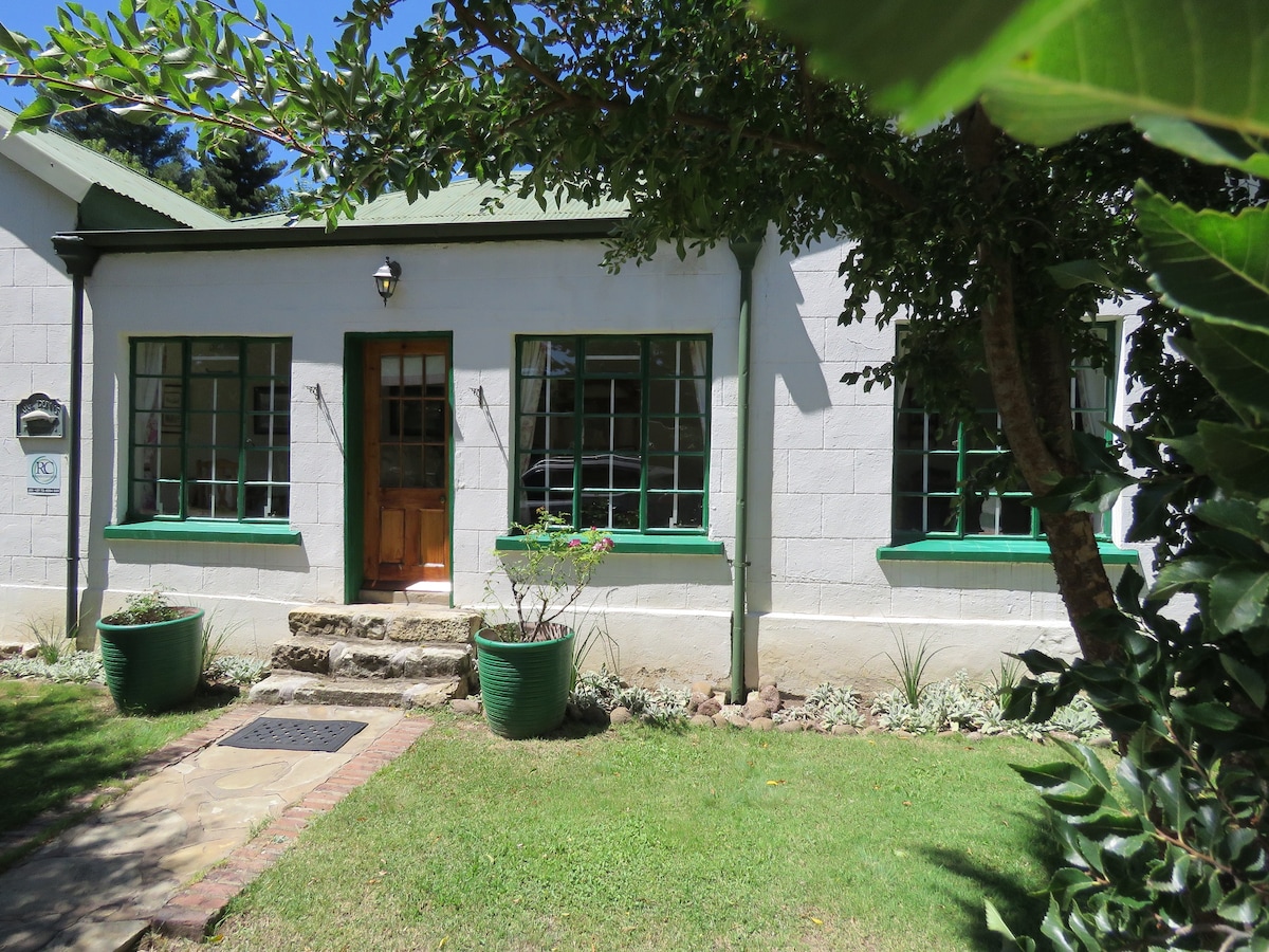 The exterior of the house features a light-colored façade, complemented by green window frames and a welcoming front door. A stone pathway leads to the entrance, surrounded by well-maintained greenery and flower planters, enhancing the inviting atmosphere of the property.