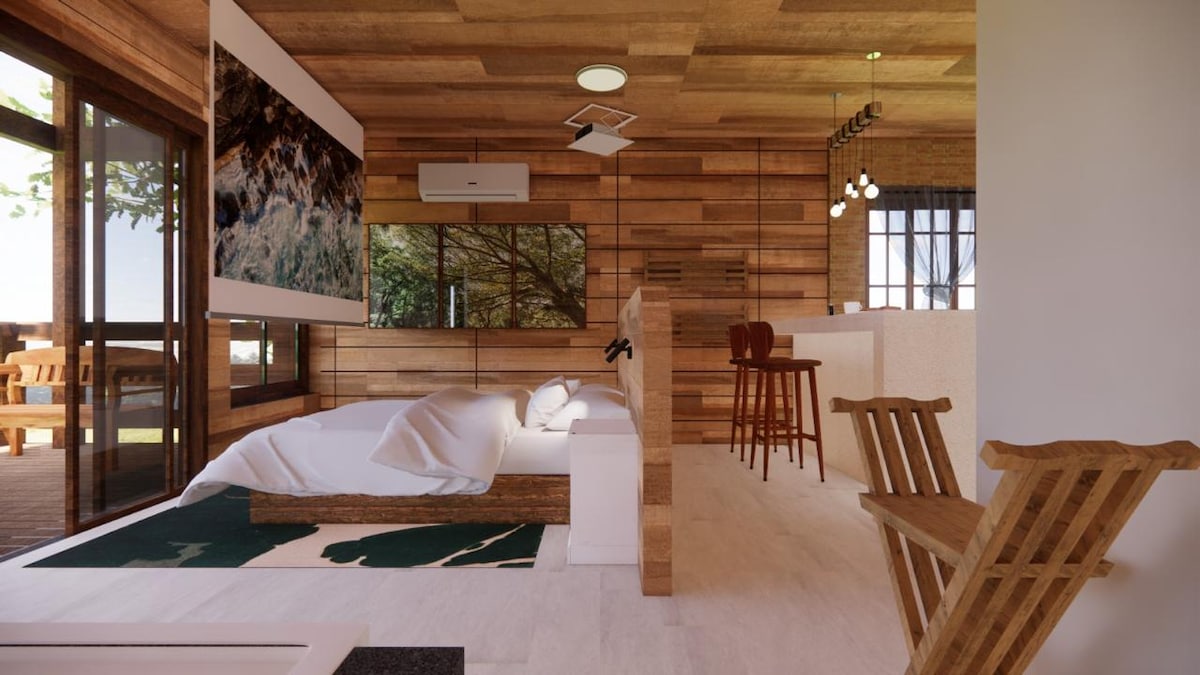 An open layout features a spacious bed adorned with a soft white coverlet. Natural wood accents create a harmonious atmosphere. A comfortable seating area is visible, along with a mini kitchen. Large windows provide ample natural light and a view of the surrounding greenery.