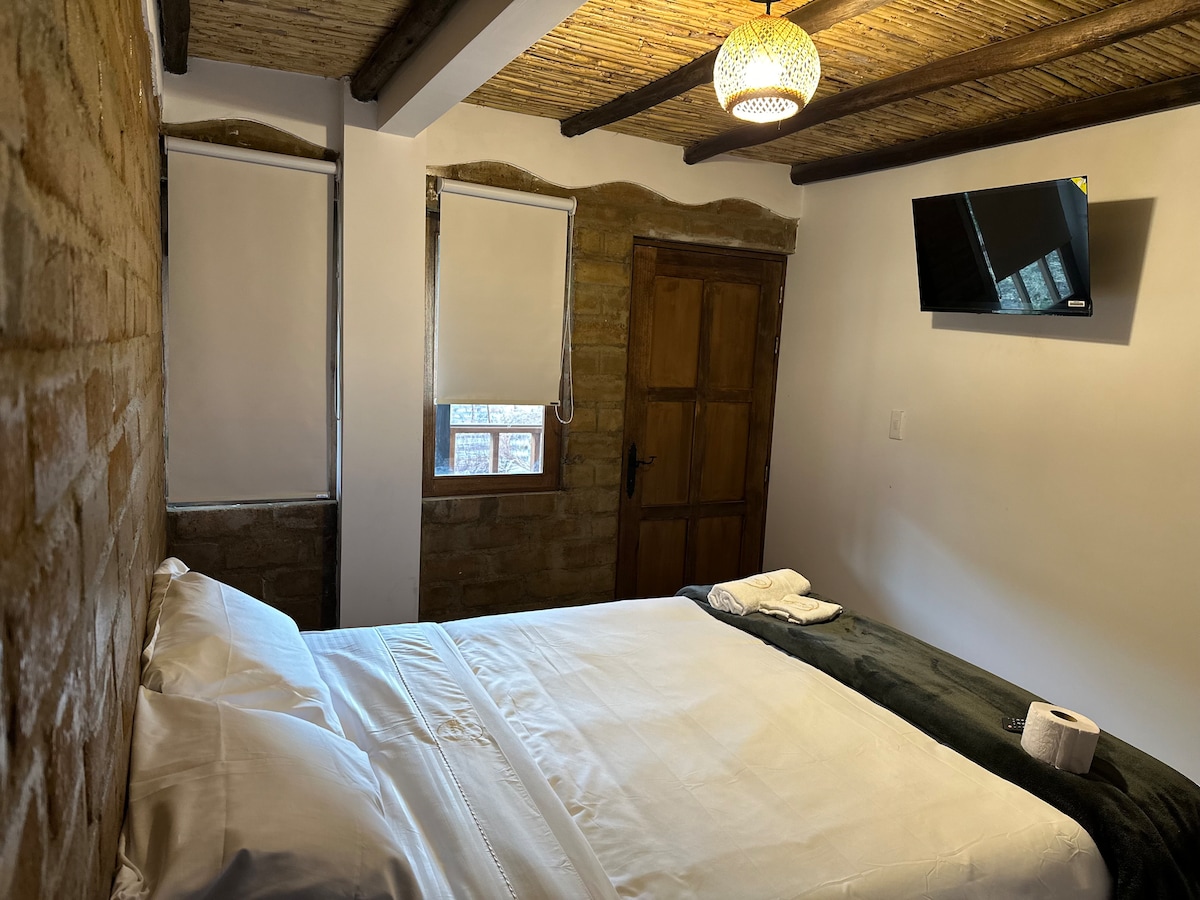 A cozy sleeping area features a spacious bed with white linens and a soft green blanket. Exposed stone walls add texture, while natural wood beams enhance the rustic feel. A small window offers natural light, complemented by rolled towels neatly placed on the bed.