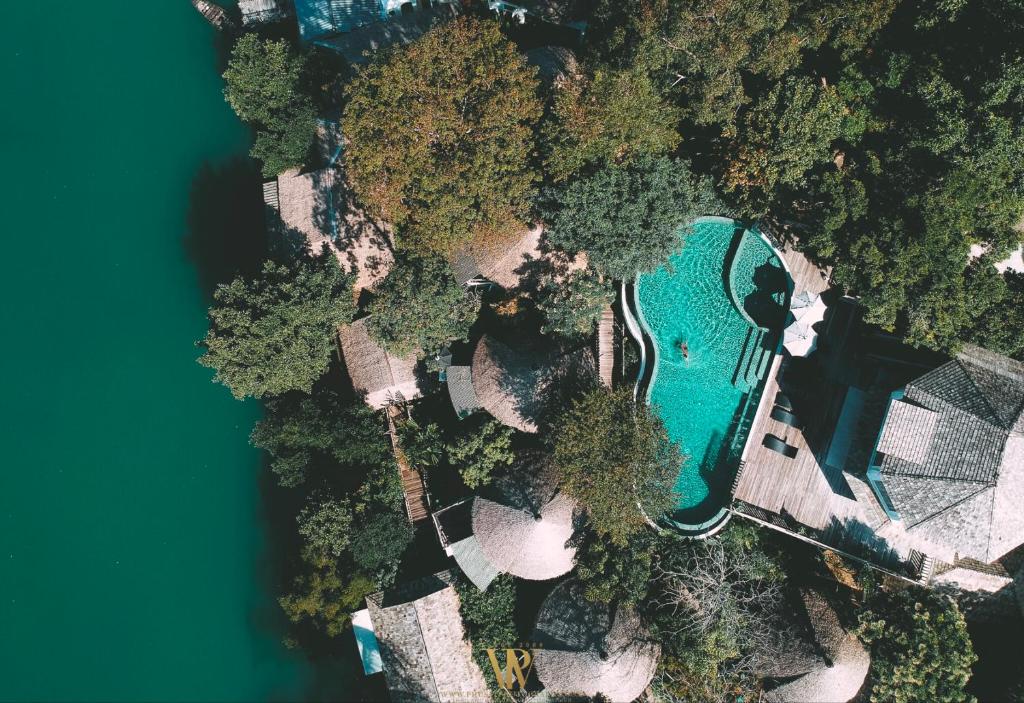 An aerial view captures a serene getaway surrounded by lush greenery. A curvy swimming pool can be seen, nestled among several thatched-roof structures. The calm water nearby reflects the vibrant foliage, creating a peaceful natural setting.