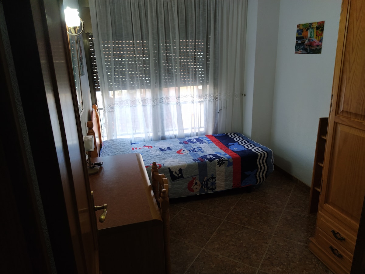 A neatly arranged single room features a bed dressed in a patterned coverlet. Natural light filters through a large window covered with sheer curtains. A wooden wardrobe and a small desk are positioned against the walls, enhancing the room's functionality.
