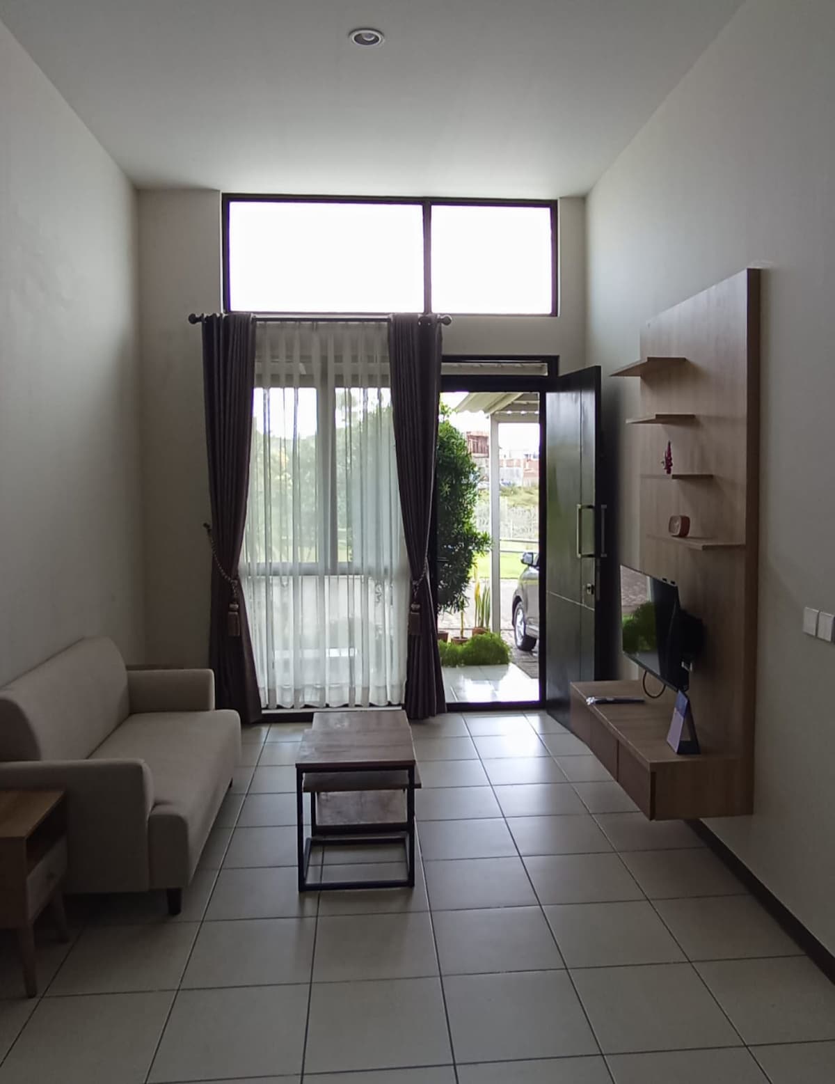 A bright living room is shown, featuring a neutral-toned sofa and a small coffee table. Large windows allow natural light to fill the space, with sheer curtains softly filtering the sunlight. A front door leads to a view of greenery outside.