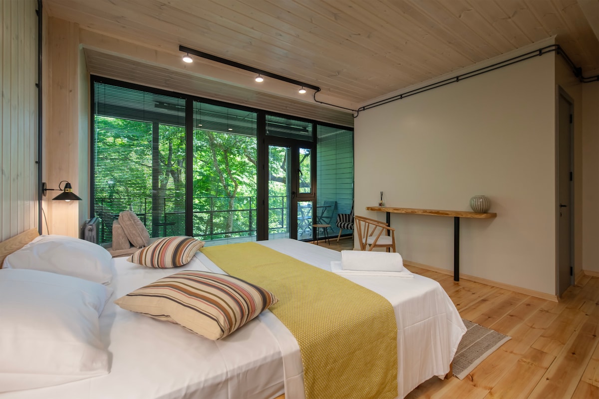 A spacious sleeping area features a large bed with striped pillows and a light yellow blanket. Natural light enters through expansive glass doors that open to a lush outdoor view. A small table and chair are positioned nearby against a wooden wall.