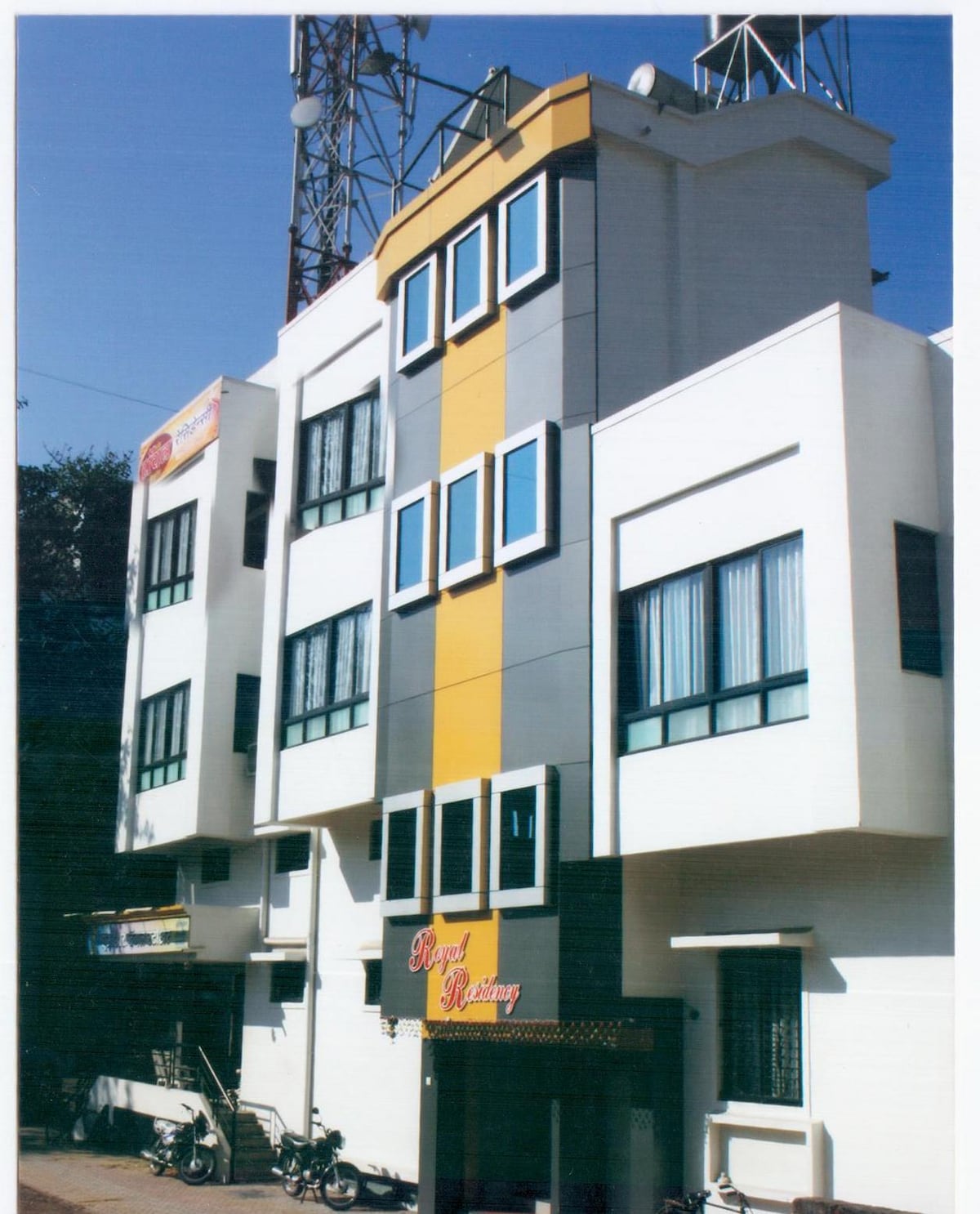An exterior view of a multi-story building reveals a modern design with a bold yellow accent running vertically down the center. Large windows provide an inviting appearance, with some windows adorned by light blue curtains. The entrance is marked by signage and has a small awning.