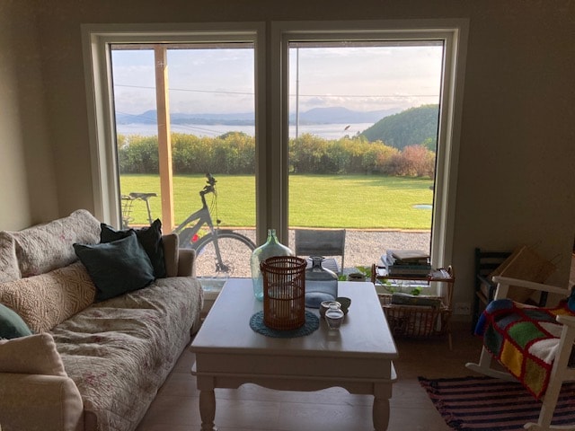 A living area is presented with a comfortable sofa and a light-colored coffee table. Two large windows frame views of the scenic outdoors, featuring a lush green lawn and water. A bicycle is visible outside, adding to the tranquil atmosphere.