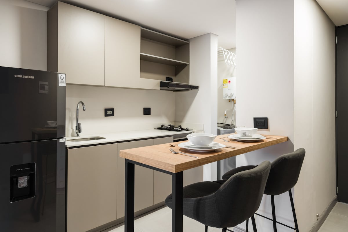 A modern kitchen area features sleek cabinets and a compact dining table with two chairs. The kitchen is equipped with a refrigerator and stove, showcasing a clean, minimalist design. Natural light illuminates the space, enhancing its functional layout.