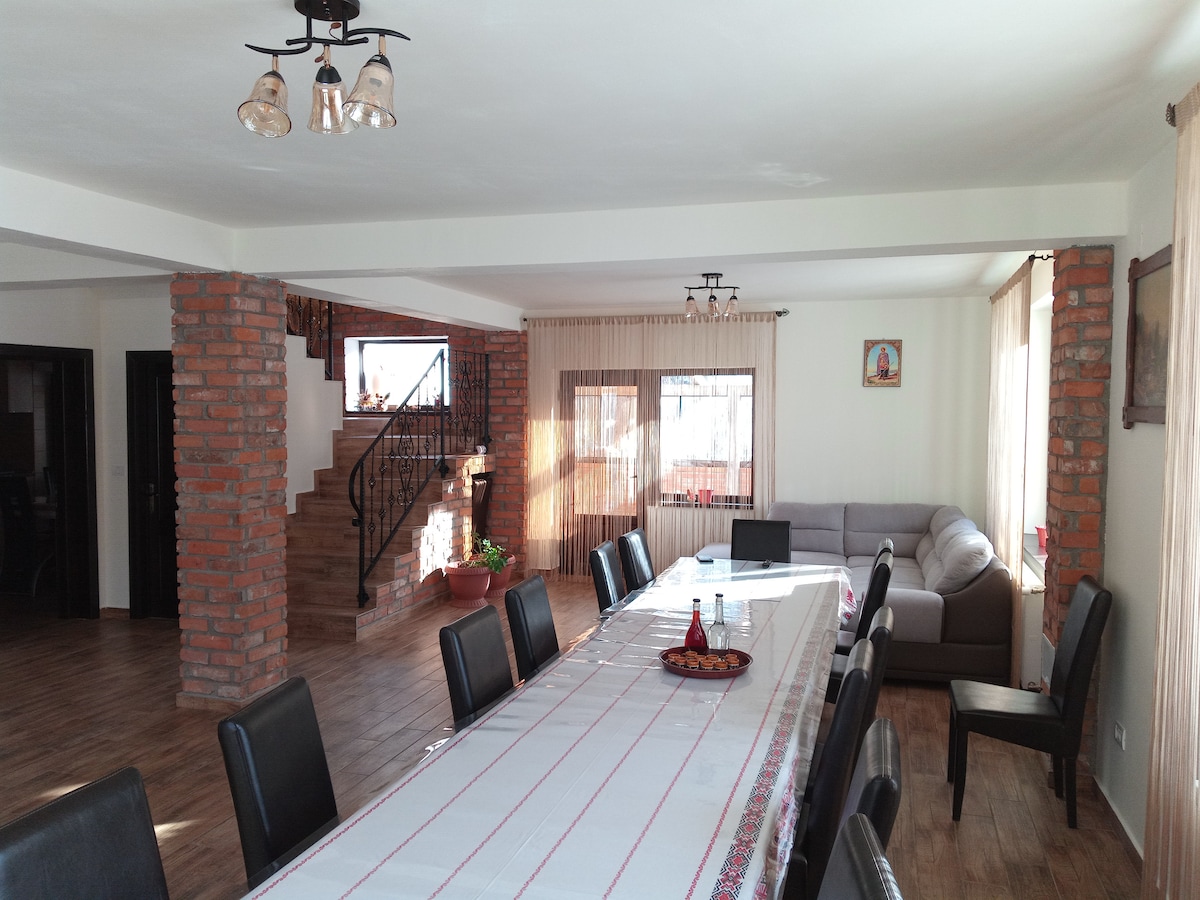 A spacious dining area features a long table surrounded by black chairs, with a decorative table runner extending the length. Large windows allow natural light to fill the room, highlighting the warm tones of the brick walls, while a comfortable sofa is visible in the background.