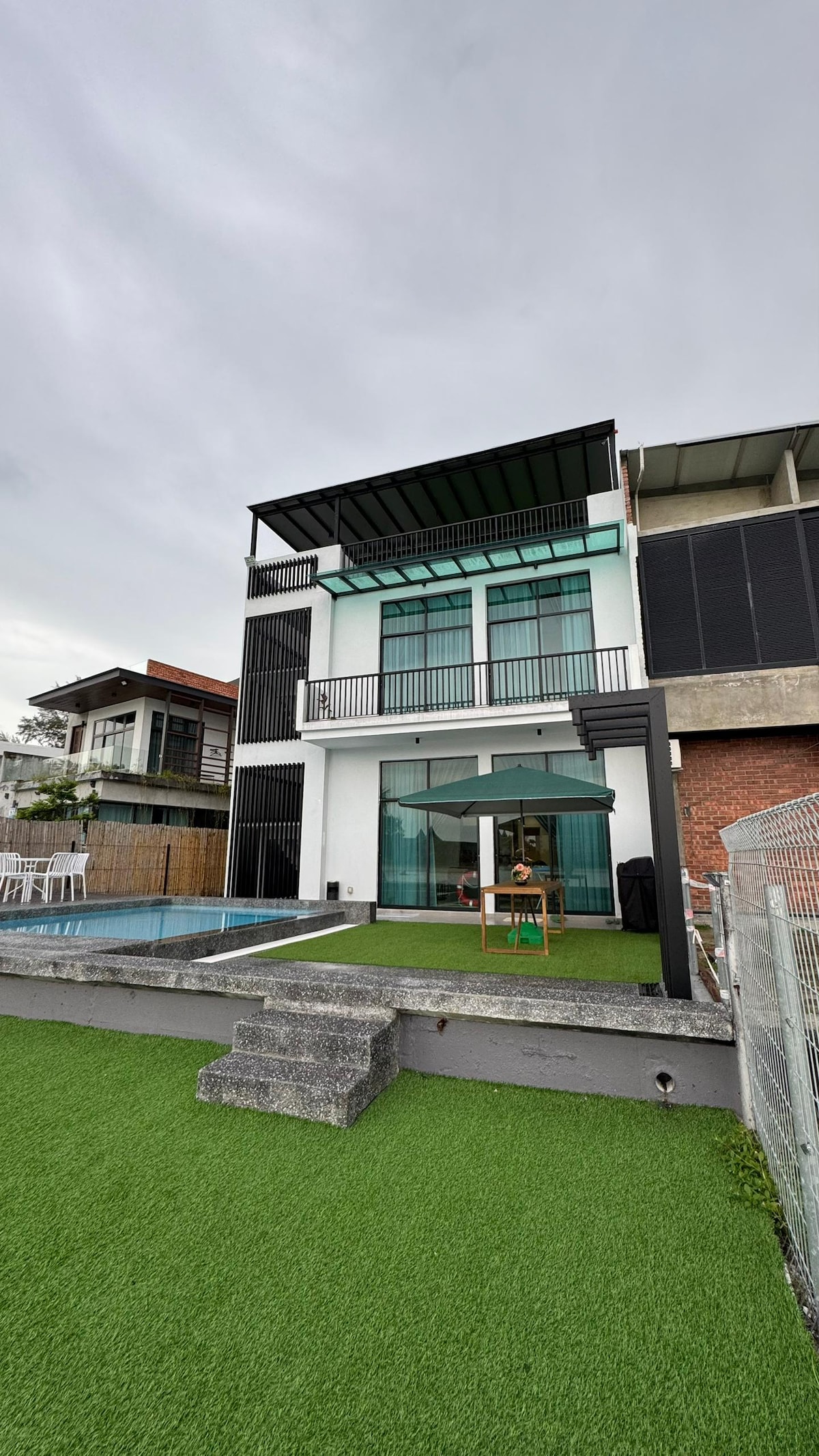 A modern villa is displayed, featuring a spacious terrace and a private pool. Lush green lawn surrounds the pool area. The villa has multiple levels, with large windows that invite natural light, and a style that blends contemporary architecture with outdoor living.