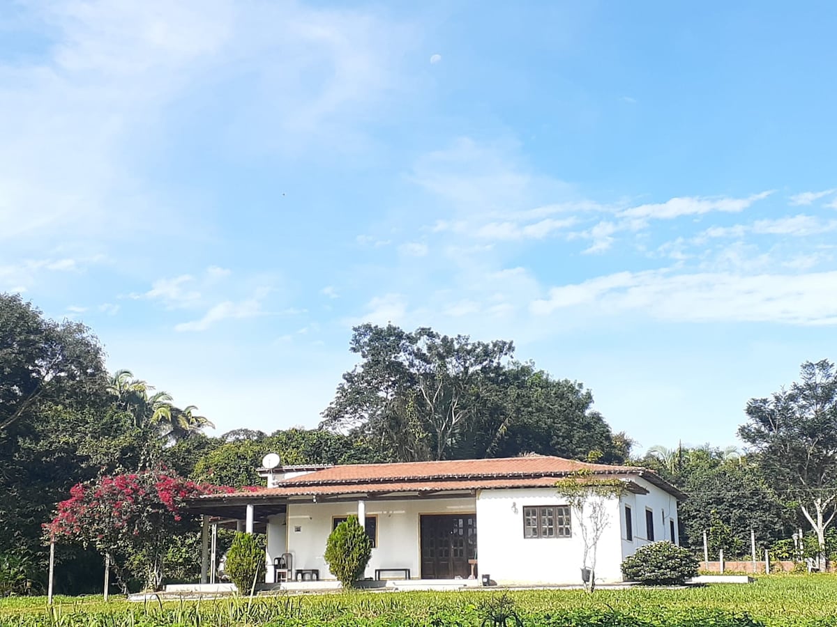 A charming countryside house is set against a clear blue sky, framed by lush greenery and vibrant flowering shrubs. The structure features a red-tiled roof and several large windows, enhancing natural light throughout the space.