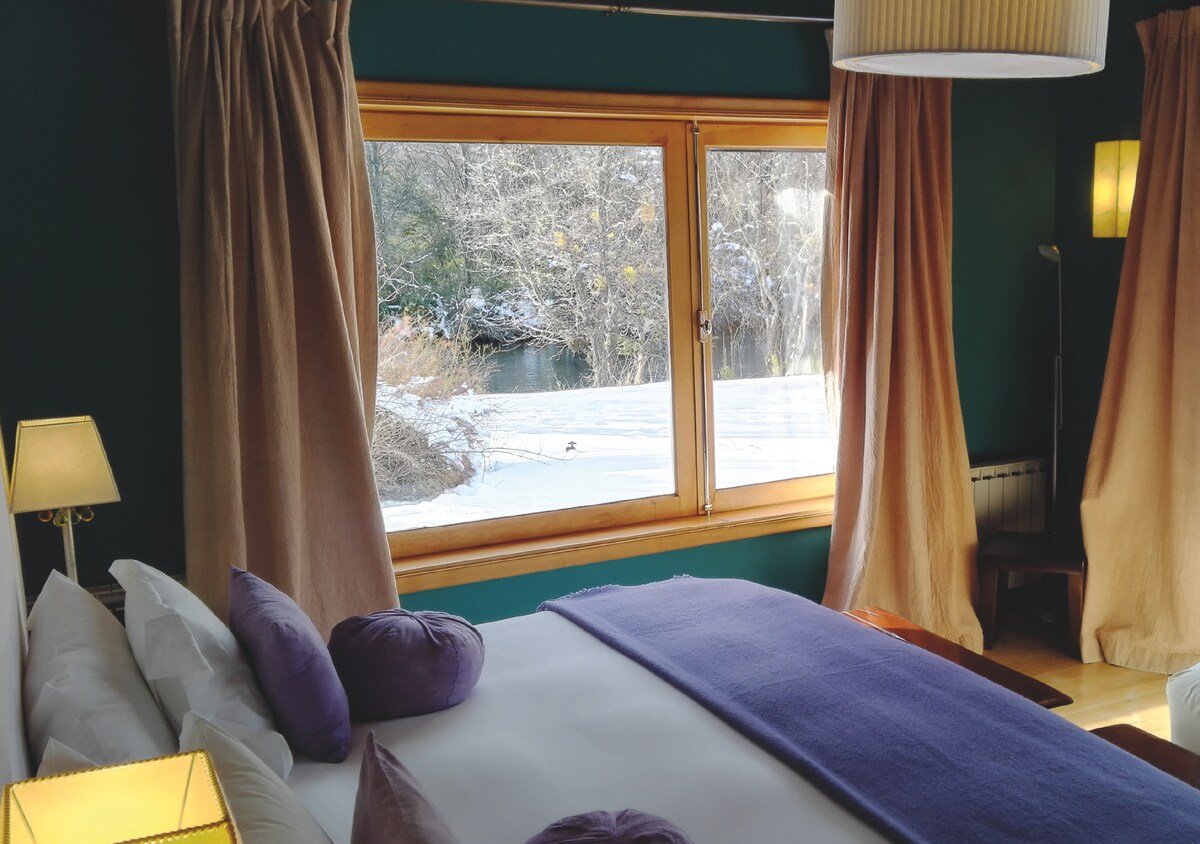 A comfortable sleeping area is presented with a spacious bed adorned with white and purple pillows. Natural light flows in from a wide window, offering views of the snowy landscape outside, framed by soft curtains in warm tones.