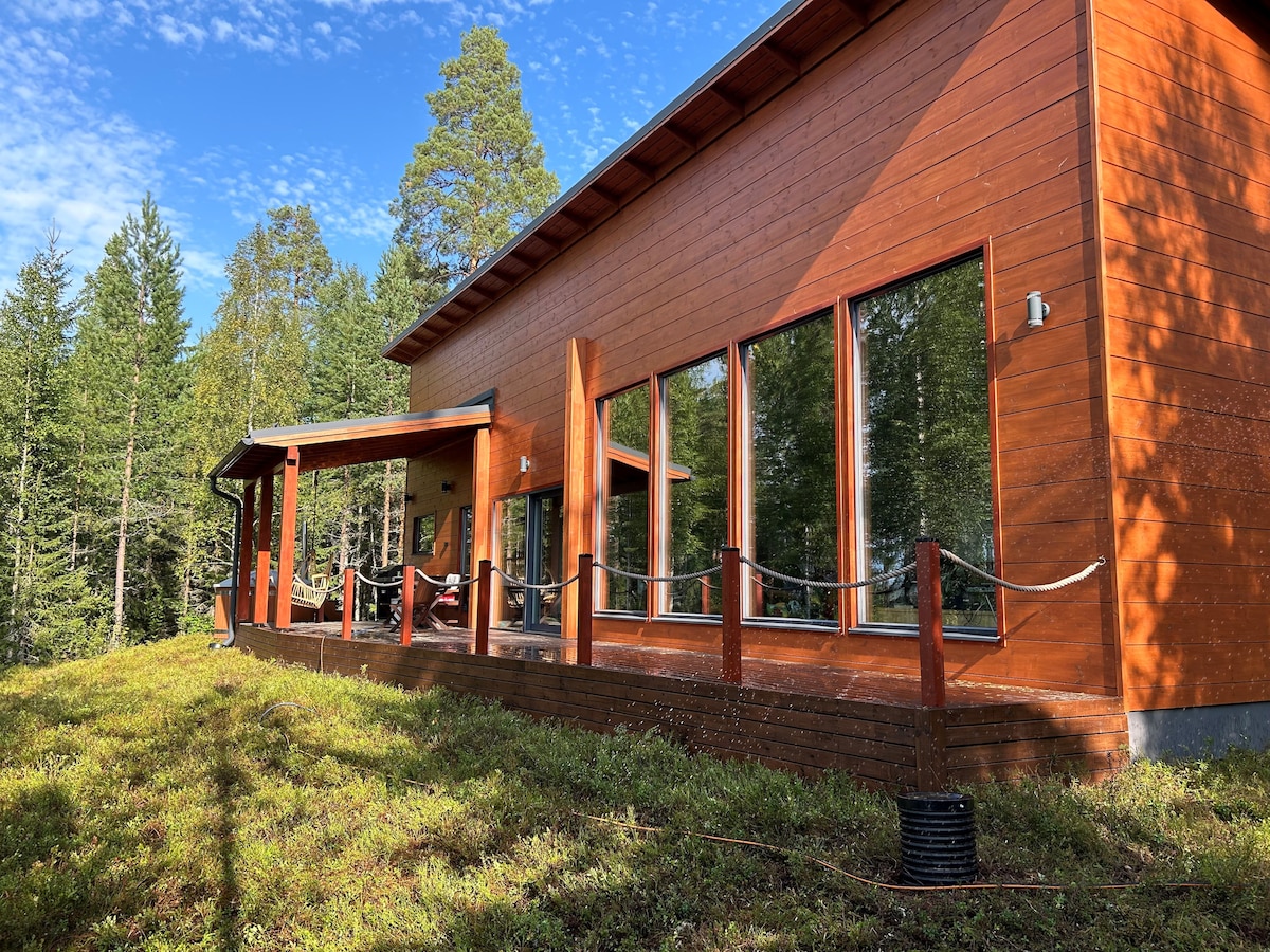 A modern wooden villa is showcased with expansive glass windows, inviting natural light. The deck is surrounded by lush greenery, providing views of the forest. Comfortable seating is visible, offering a serene outdoor space.