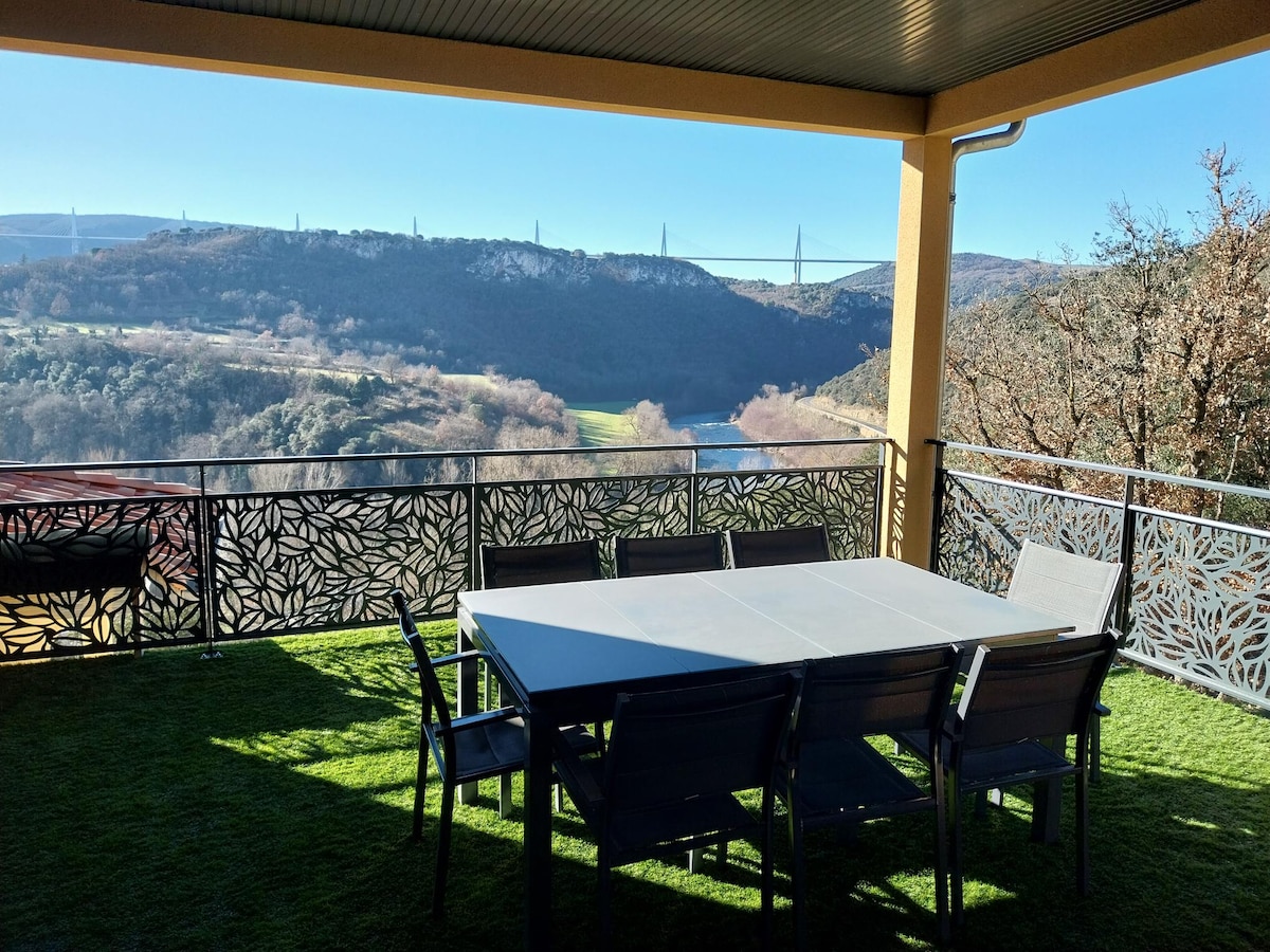 A covered terrace features a dining table surrounded by chairs, offering a view of rolling hills and the Millau Viaduct in the distance. The area is adorned with decorative railings and lush green grass underfoot.