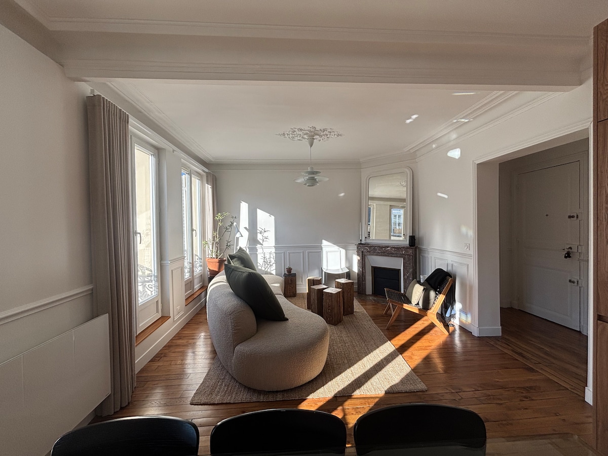 A spacious living area is highlighted by large windows allowing natural light to flow in. A unique curved sofa is positioned on a textured rug, accompanied by wooden stools. A stylish armchair and a fireplace enhance the comfort of this well-designed space.