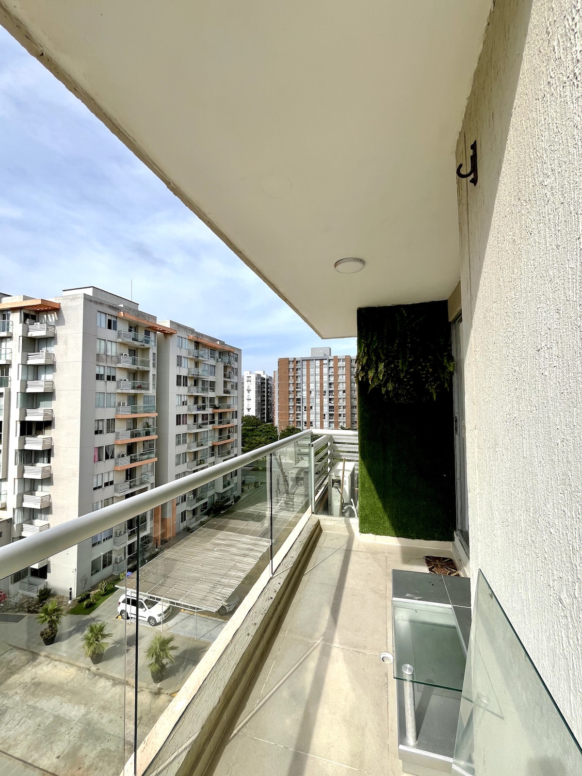 A spacious balcony is shown, featuring glass railings that provide an unobstructed view of neighboring buildings and the surrounding area. The floor is tiled, and a small glass table is visible. Greenery adorns the wall, adding a touch of nature to the urban setting.