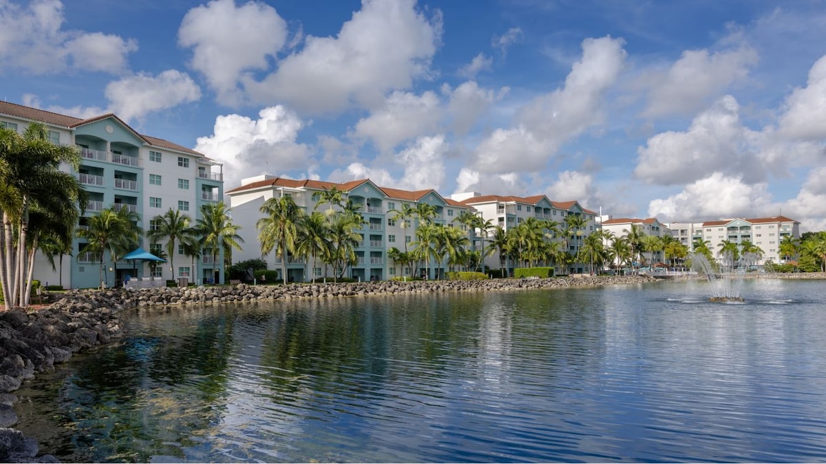 A tranquil waterfront view shows the resort's modern buildings lined along the lake, surrounded by tropical palm trees. The clear blue sky and puffy clouds reflect gently on the water's surface, while a fountain creates a serene focal point in the background.
