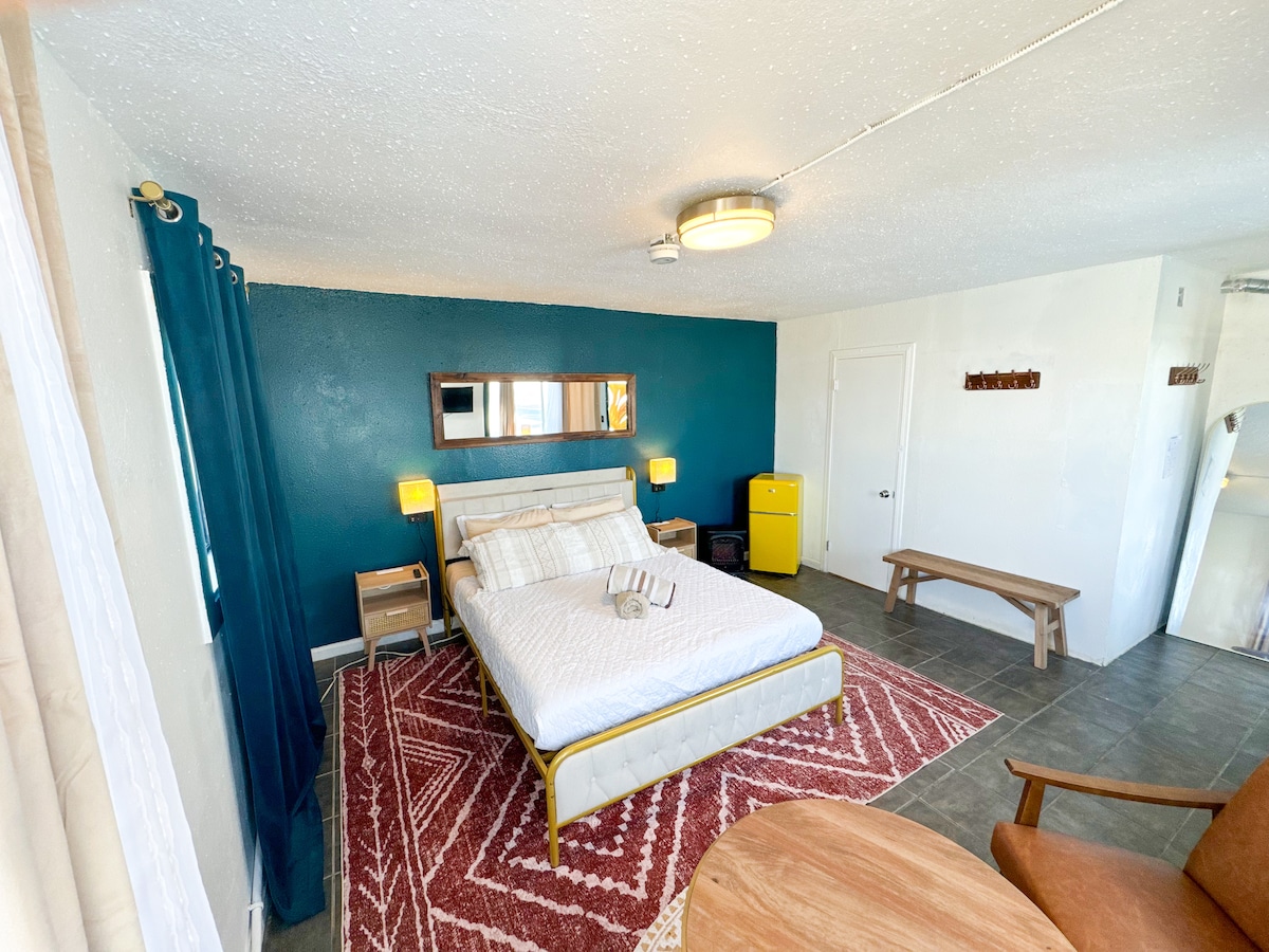 A spacious room is shown, featuring a large bed with crisp white linens and a decorative throw. A teal accent wall contrasts with neutral furnishings, including two bedside lamps, a mirrored wall, and a small yellow refrigerator. A wooden table and chair are positioned nearby.