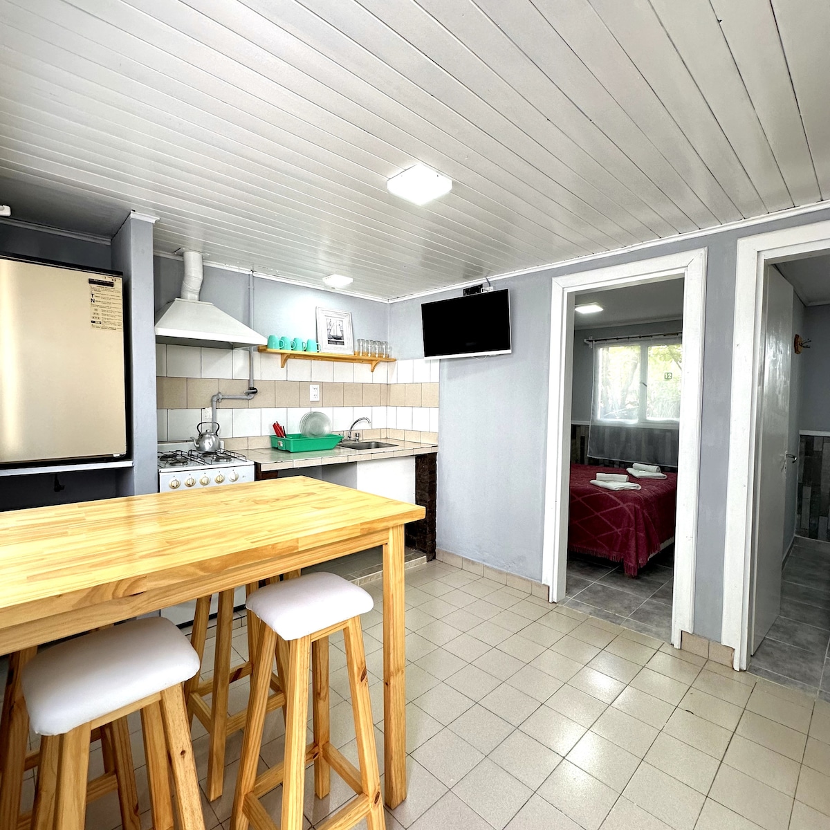 A functional kitchen area features a wooden bar table with three stools, accompanied by a stove and overhead range hood. The space is illuminated by ceiling lights, and a television is mounted on the wall. A doorway leads to a separate sleeping area in the background.