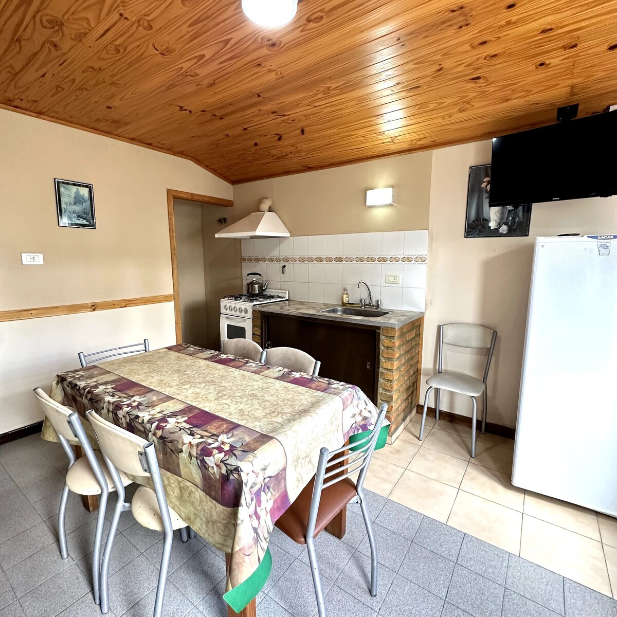 A spacious kitchen area features a large dining table set for six, topped with a floral tablecloth. A well-equipped kitchen includes a white refrigerator, a stove, and ample counter space. Wooden ceiling beams add a natural touch to the neutral-toned walls.