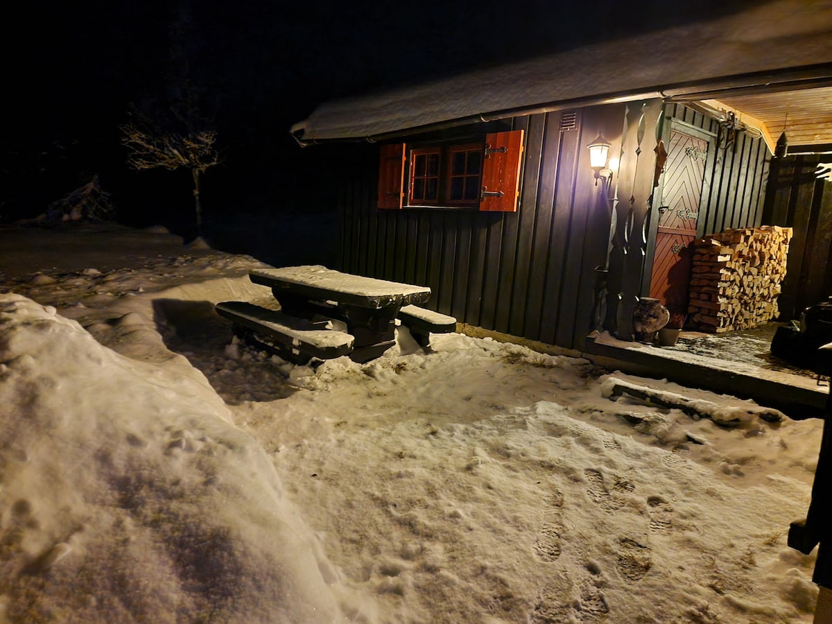 A rustic cabin exterior is illuminated by a soft lantern light, featuring a wooden picnic table and benches in the foreground. Snow blankets the ground, accentuating the dark walls and the warmly colored window shutters. Stacked firewood adds to the cozy ambiance.