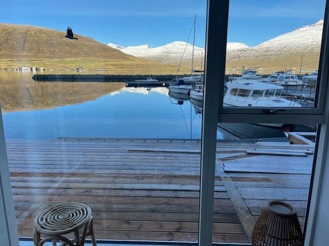 The image captures a serene view from the interior, showcasing a calm marina with boats moored alongside a wooden dock. Snow-capped hills rise in the background under a clear blue sky, reflecting the tranquil atmosphere of the environment.