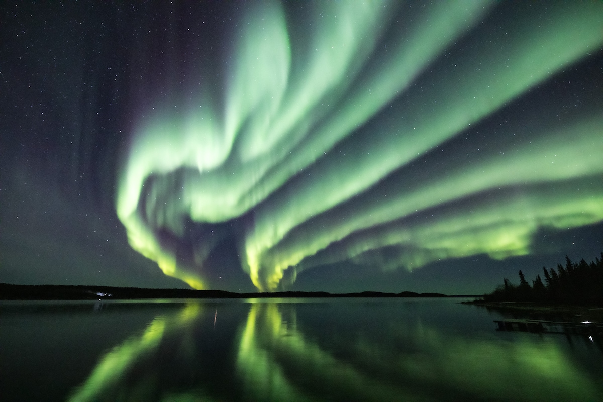 A stunning view of the Northern Lights illuminates the night sky with vibrant green and subtle hints of yellow. The colorful display reflects off the calm surface of the lake, framed by a silhouette of distant trees along the shoreline.