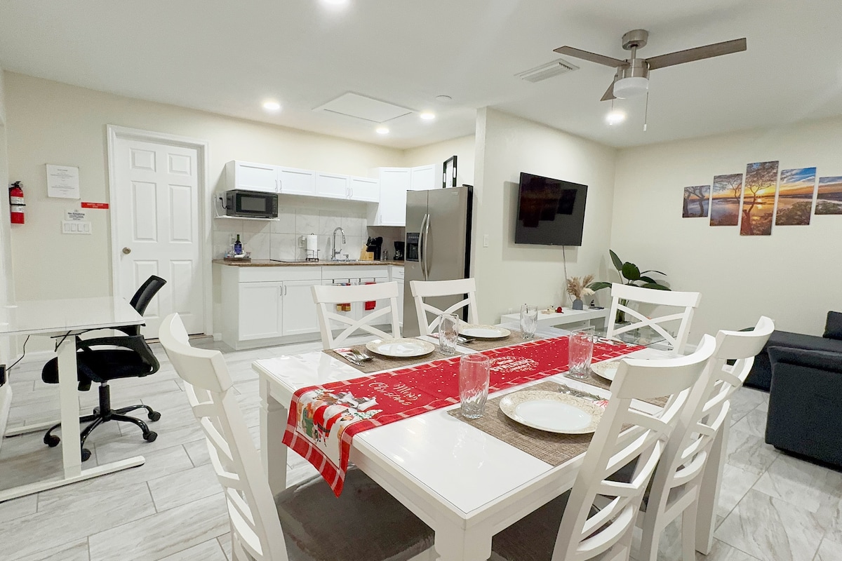 The open-plan kitchen and dining area features a large white table with six chairs, set for a meal with plates and glassware. A stainless steel refrigerator and microwave are visible, along with a comfortable sofa and a wall-mounted TV. Soft lighting enhances the clean and functional design.