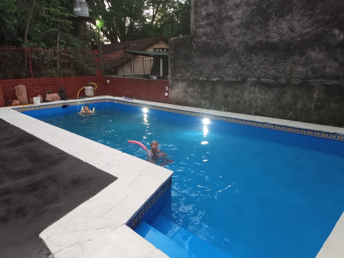 A well-maintained swimming pool occupies the center of the image, with clear blue water reflecting light from above. The pool is bordered by white concrete, providing a sharp contrast. Lush greenery and a brick wall are visible in the background, contributing to a peaceful setting.
