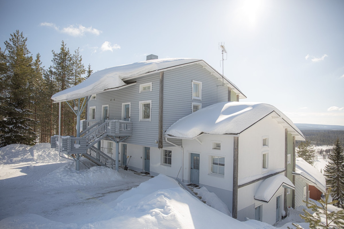 A snow-covered exterior of a multi-story building is visible, showcasing a light-colored wooden façade. Steps lead to the main entrance, while trees frame the property. The sky is clear and bright, complementing the serene winter landscape.