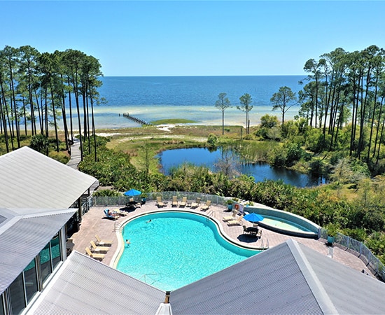 An aerial view captures a spacious pool surrounded by lounge chairs, with vibrant blue waters reflecting the clear sky. In the background, the serene beach meets lush greenery, leading to calm waters, while tall trees line the edges of the scene.