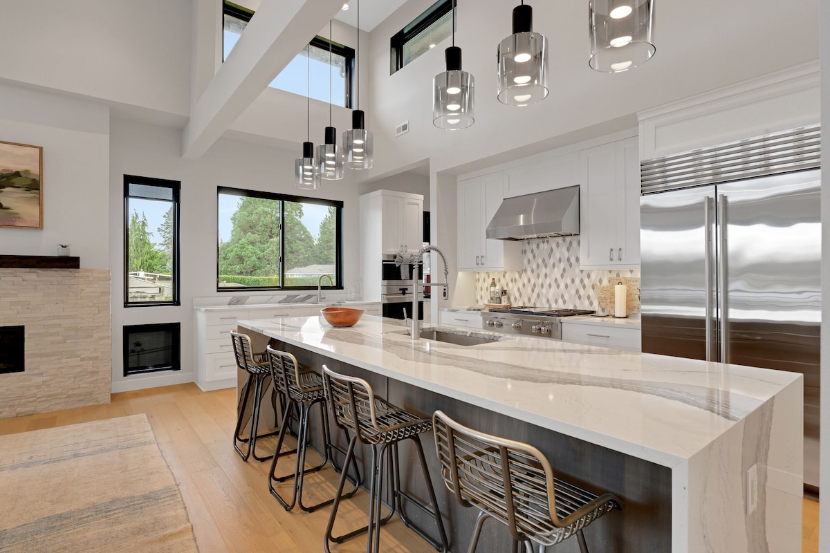 A modern kitchen features a spacious island with bar seating for four, complemented by sleek pendant lights overhead. Stainless steel appliances and white cabinetry create a clean aesthetic, while large windows invite natural light and provide views of the surrounding greenery.