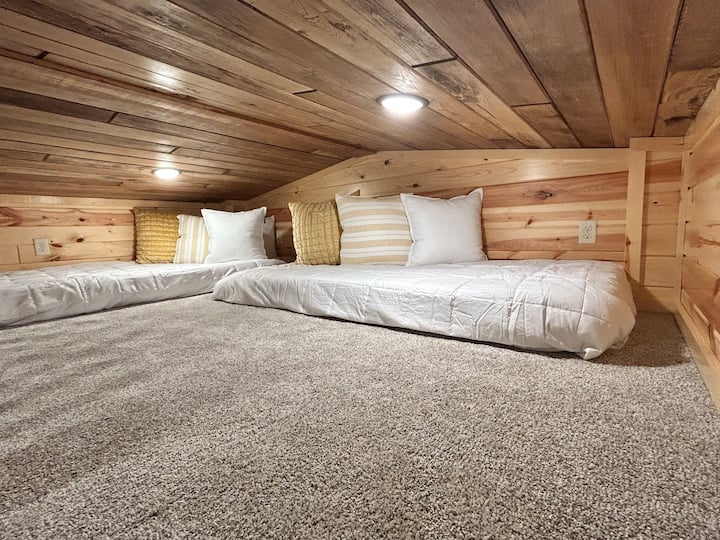 Cozy loft sleeping area accessed by a ladder, featuring 2 foam twin beds with fresh linens. The loft has a warm wood finish, soft carpeting, and built-in lighting for a snug, cabin-style feel. 2 portable fans are available in the closet below.