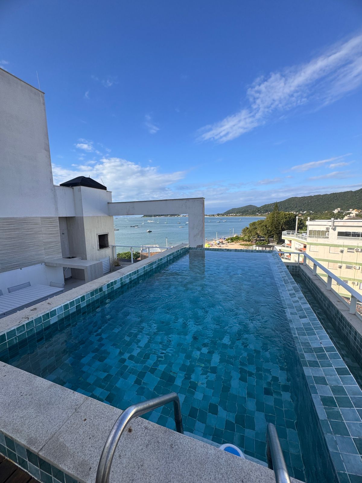 An infinity edge pool is positioned overlooking a scenic coastline. The pool features tiled steps leading into the water, surrounded by a spacious deck. A clear sky with scattered clouds and a view of distant hills and boats is visible in the background.