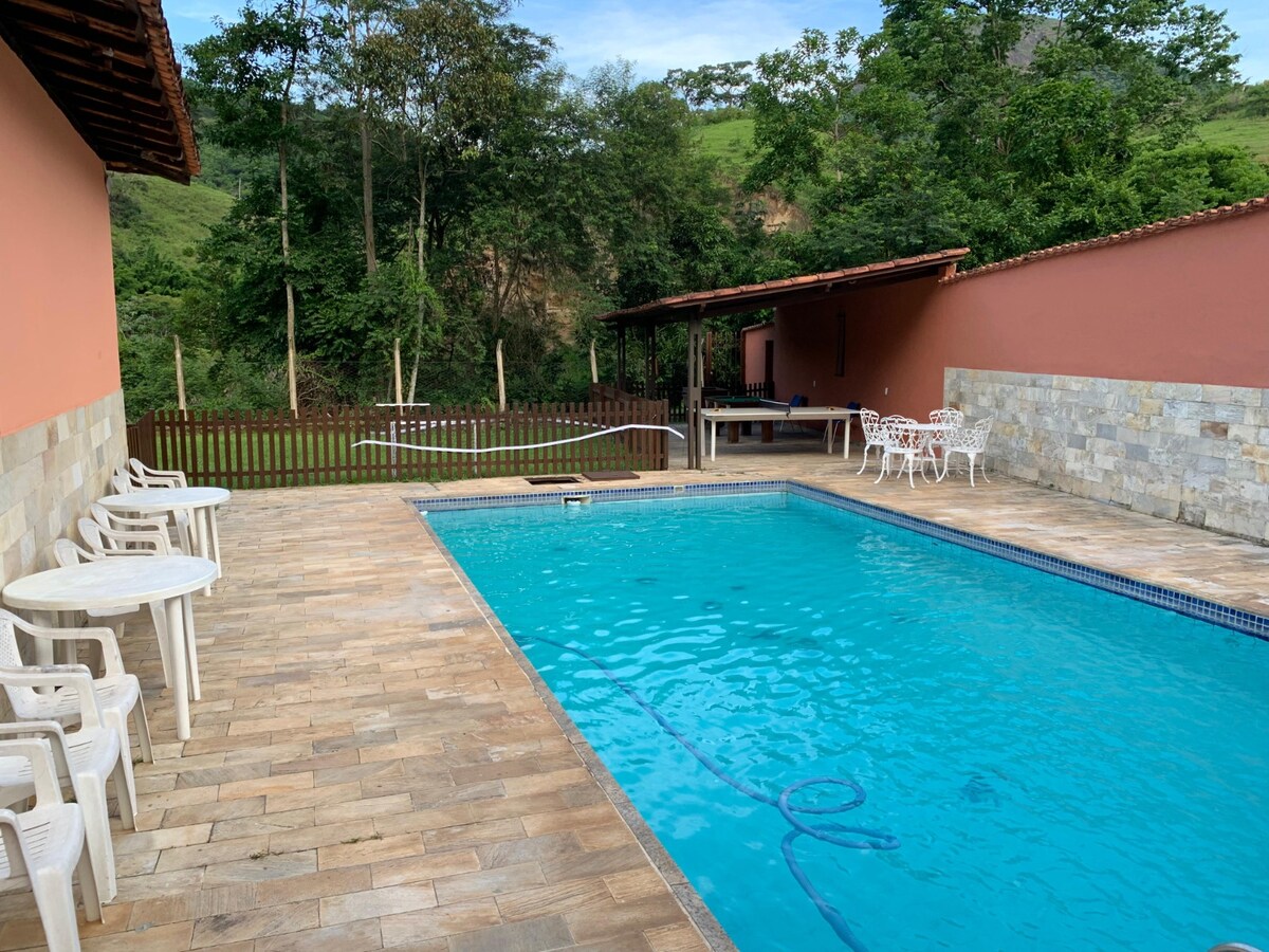 A vibrant outdoor area features a clear swimming pool surrounded by stone tiles. White seating chairs are arranged on one side, with a shaded area visible beyond. Lush greenery and trees provide a natural backdrop, enhancing the serene environment.