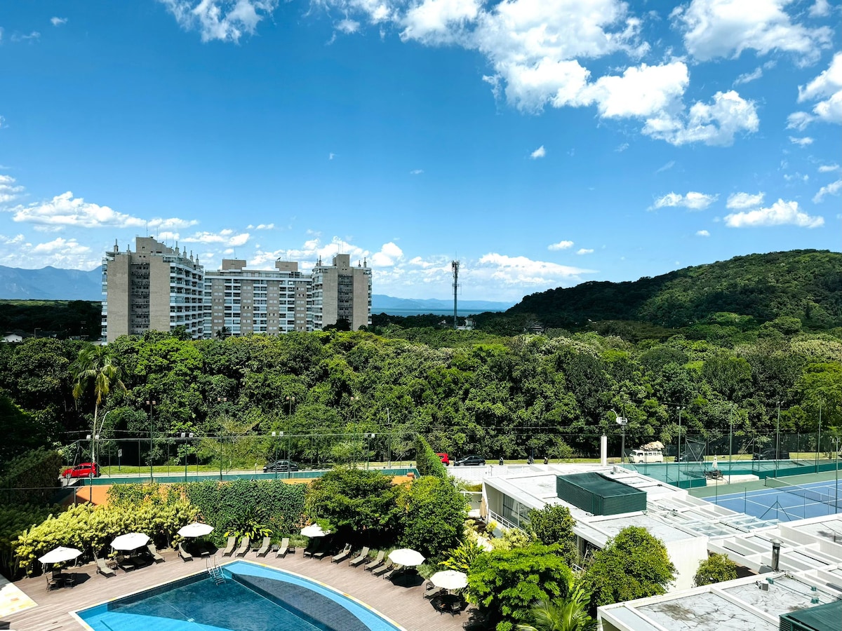 A panoramic view captures the resort's inviting swimming pool and expansive greenery, framed by distant mountains under a clear blue sky. The scene features lounge chairs arranged around the pool, emphasizing an atmosphere of relaxation and leisure.