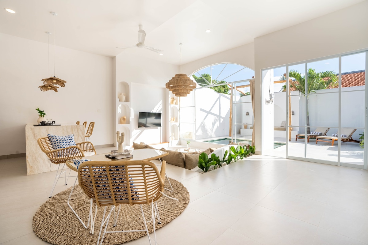 An airy living space features a round dining table surrounded by woven chairs. A large sofa with cushions is positioned nearby. Light pours in through glass doors, revealing a private pool and outdoor area, complemented by tropical plants and modern decor.