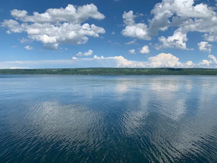 The image captures a serene view of Seneca Lake, showcasing calm waters reflecting a vibrant sky filled with fluffy clouds. The distant shoreline is lined with greenery, contributing to the peaceful atmosphere of the lakeside setting.