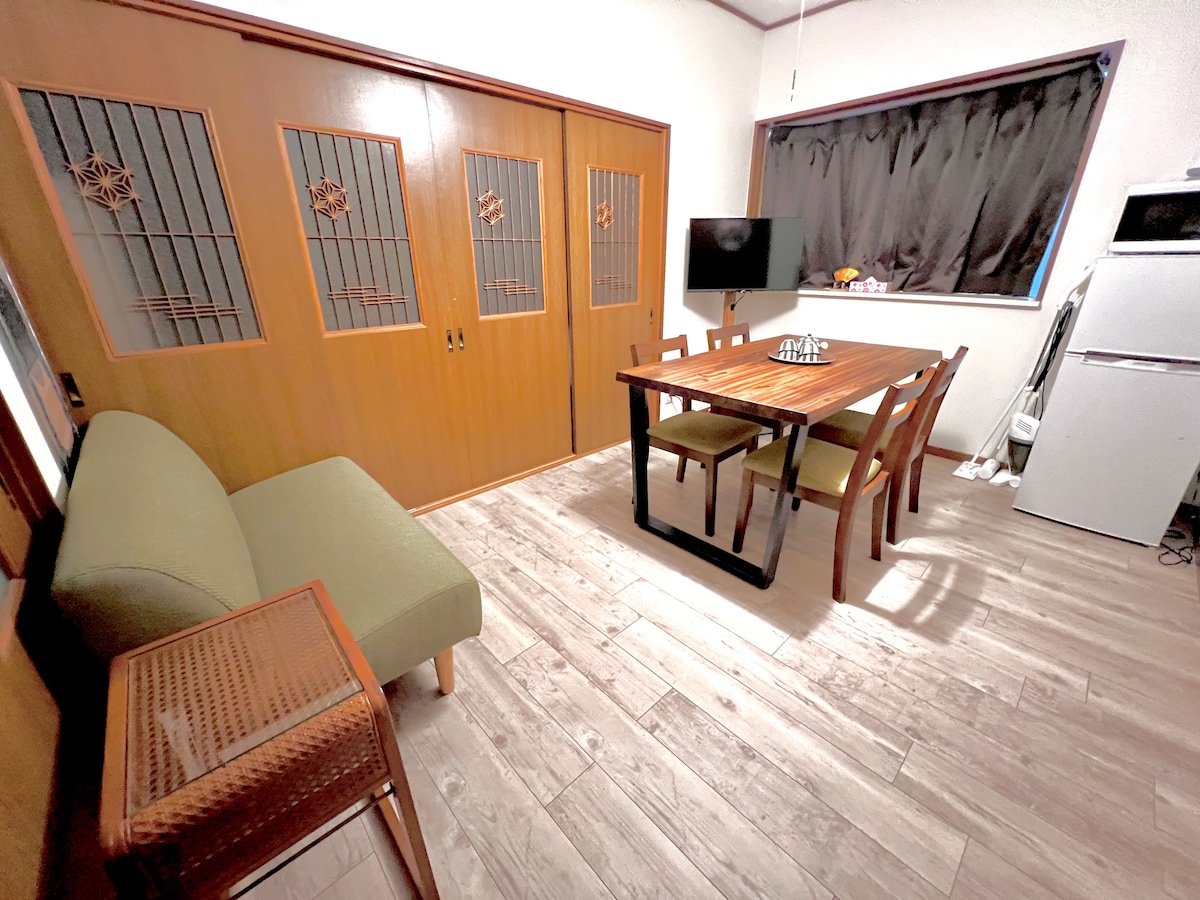 A living area is shown, featuring a dining table for four and a small green sofa. A television is positioned near the window, which is dressed with dark curtains. Wooden doors with decorative panels add to the character of the space.