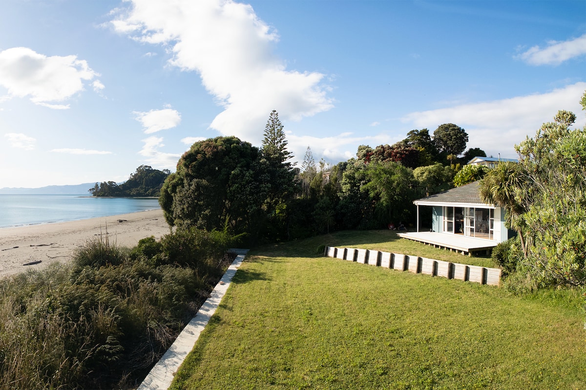 A beachside property is framed by lush greenery and a sandy shoreline. A spacious lawn leads to a wooden deck, offering views of the calm water and distant hills under a clear sky. The house features large windows that bring in natural light.