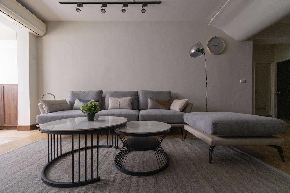 A cozy living area is furnished with a gray sectional sofa and a stylish chaise lounge. Two coffee tables, one round and one oval, are positioned on a textured rug. Light from ceiling spotlights creates a welcoming ambiance, enhancing the room's modern aesthetic.