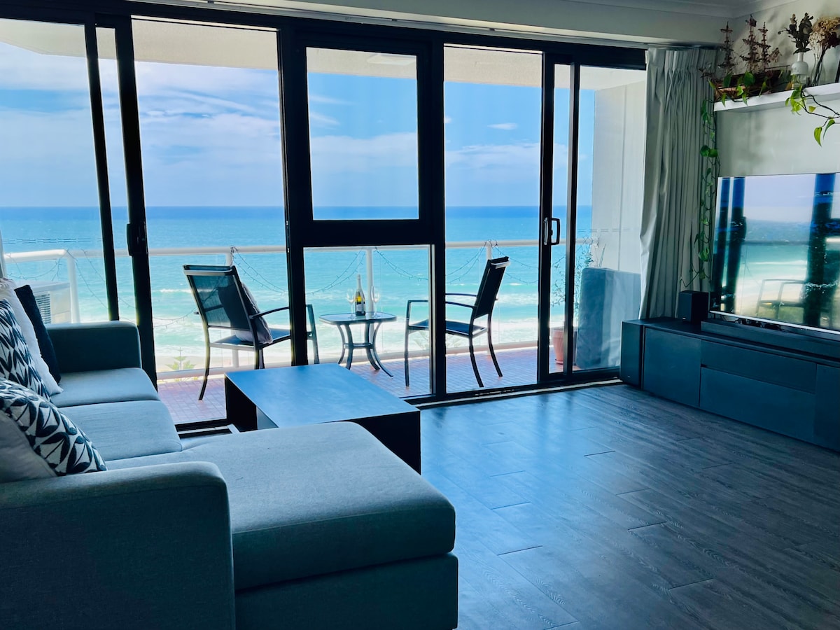 Floor-to-ceiling glass doors frame panoramic ocean views, allowing natural light to fill the open living area. A comfortable sofa faces a modern media console, while a small balcony seating arrangement of two chairs and a table offers a perfect spot for enjoying the seaside scenery.